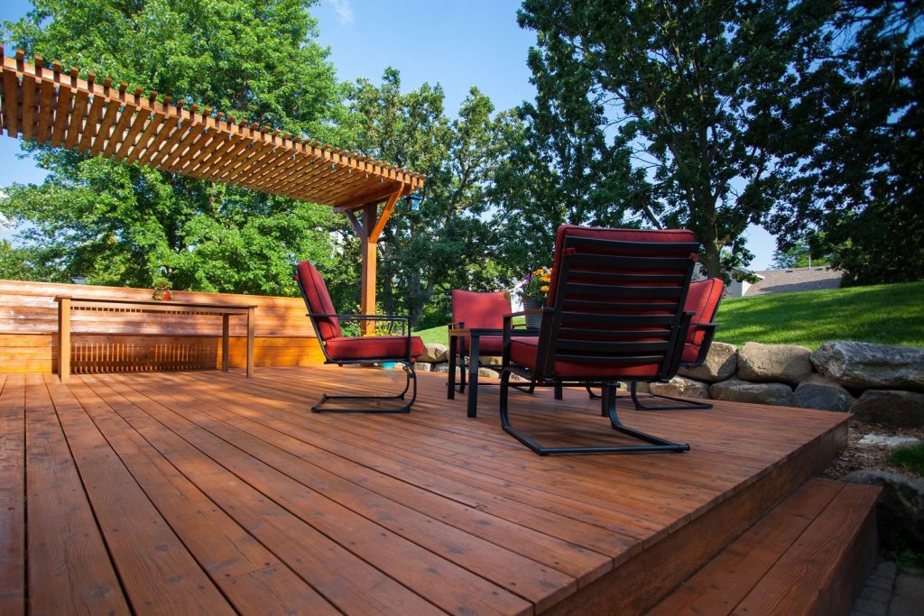 Wooden patio deck with red cushioned chairs, a table, and a pergola, surrounded by greenery and trees.