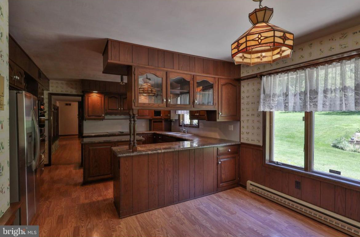 Kitchen with dark wood cabinets, a pendant light, large window with lace curtains, and wood floors.