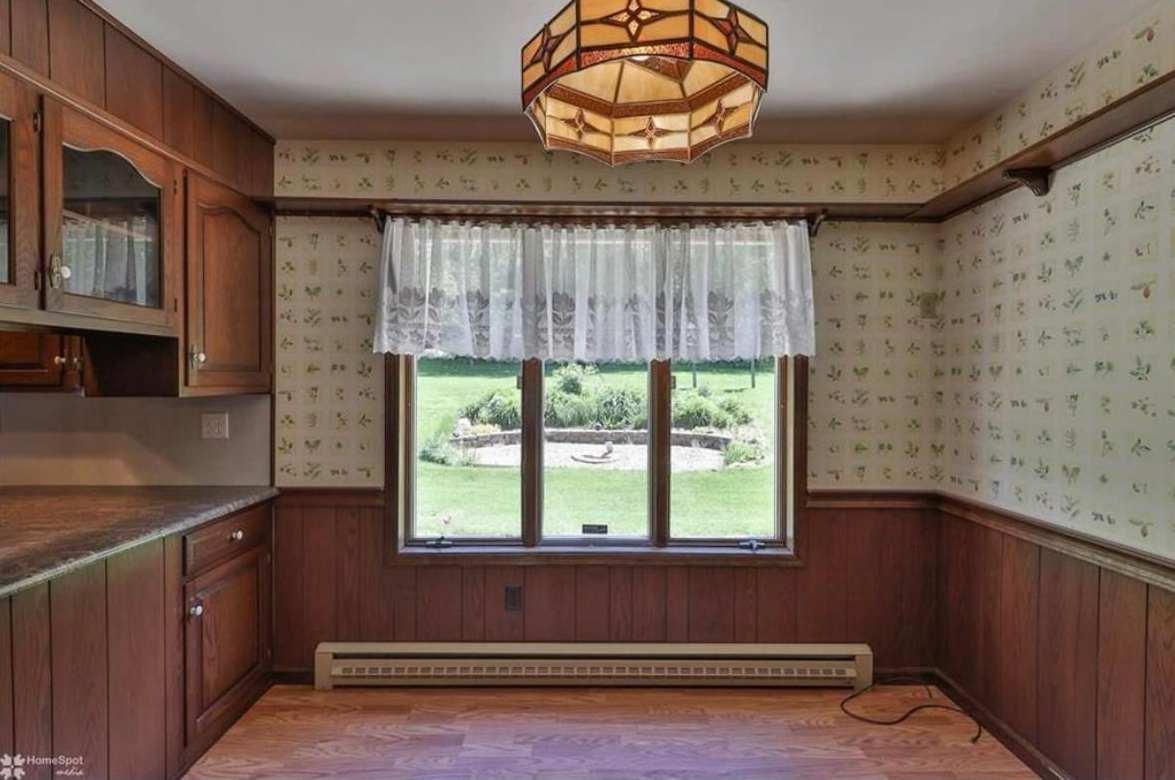 A vintage kitchen with wood cabinets, floral wallpaper, lace curtains, and a geometric pendant light above wood flooring.