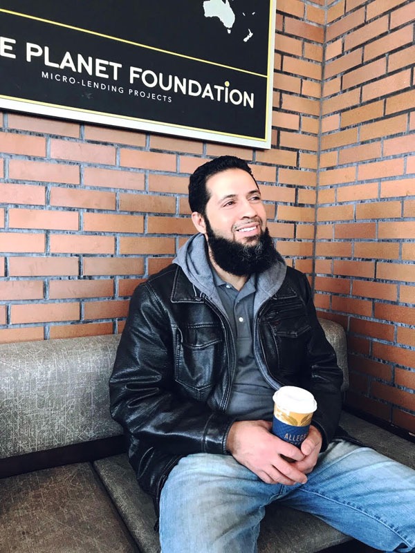Man with a beard sitting on a bench, holding a coffee cup, smiling, with a brick wall and foundation sign behind him.